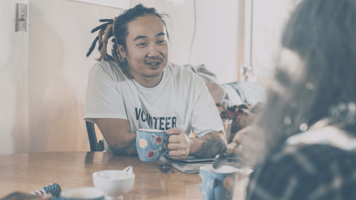 A male presenting person smiling and talking at a kitchen table with another person, holding coffee
