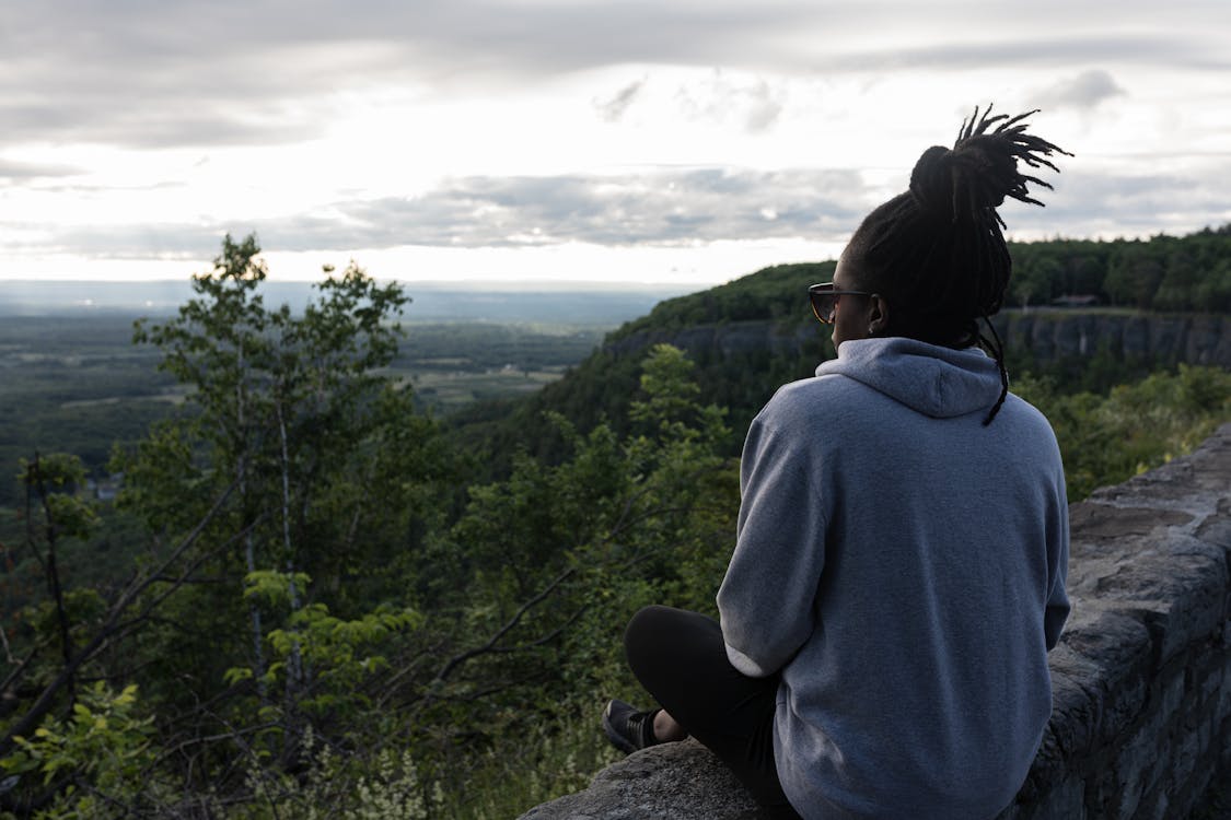 A young Black person sits on a ledge and looks out over a valley landscape