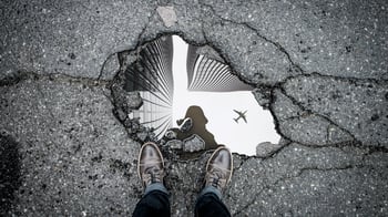 Photo of a man's shoes in front of a puddle reflecting up at the sky.