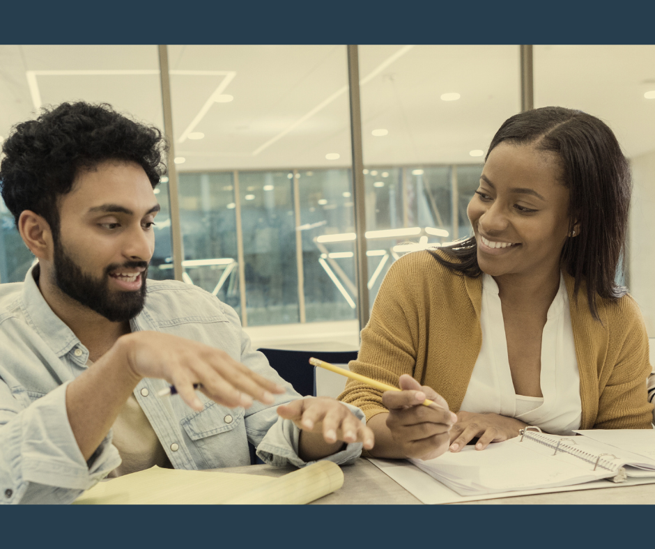 A male presenting person and a female presenting person sitting at a table doing work together, smiling