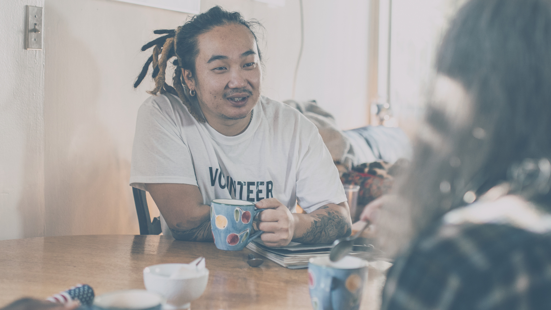 A male presenting person smiling and talking at a kitchen table with another person, holding coffee